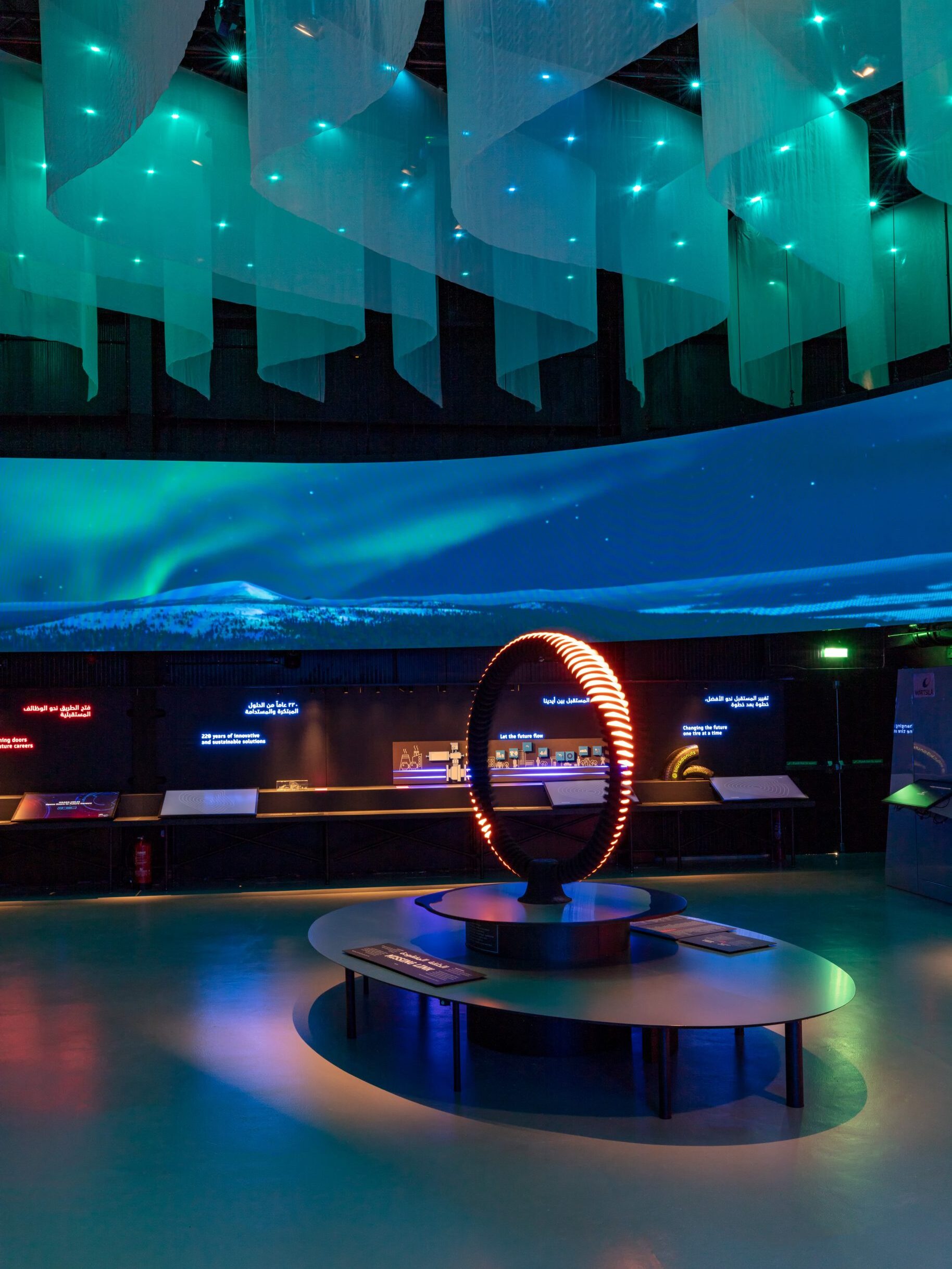 Pavilion hall showing the northern lights over a snowy landscape on a wall-sized screen, with an illuminated ring sculpture on a central plinth